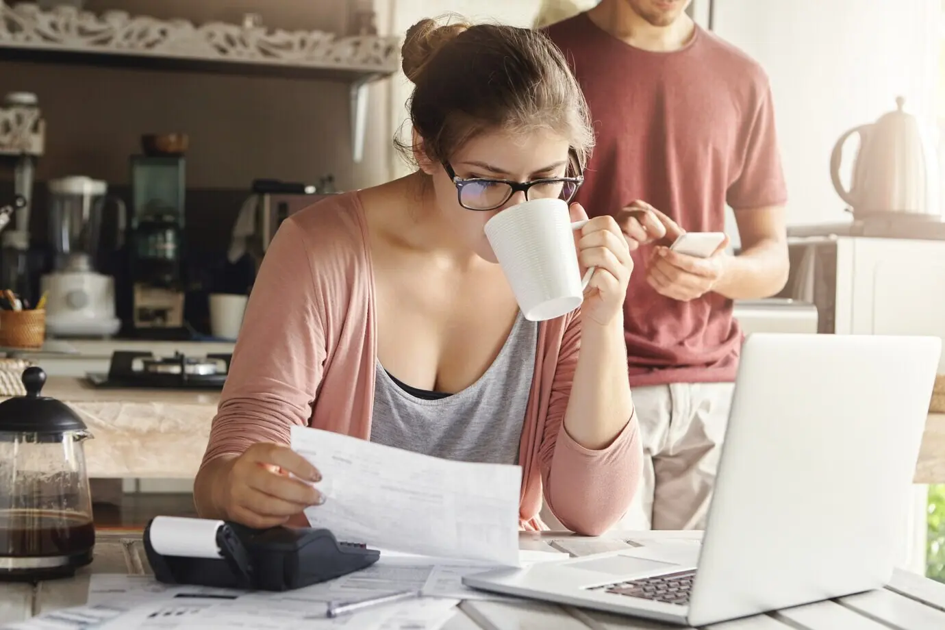 Attraktive, ernsthafte Frau mit Brille trinkt Kaffee und prüft ein Dokument in ihren Händen; sie verwaltet das Familienbudget und erledigt Papierkram am Küchentisch, auf dem sich ein Stapel Rechnungen, ein Laptop und ein Taschenrechner befinden.